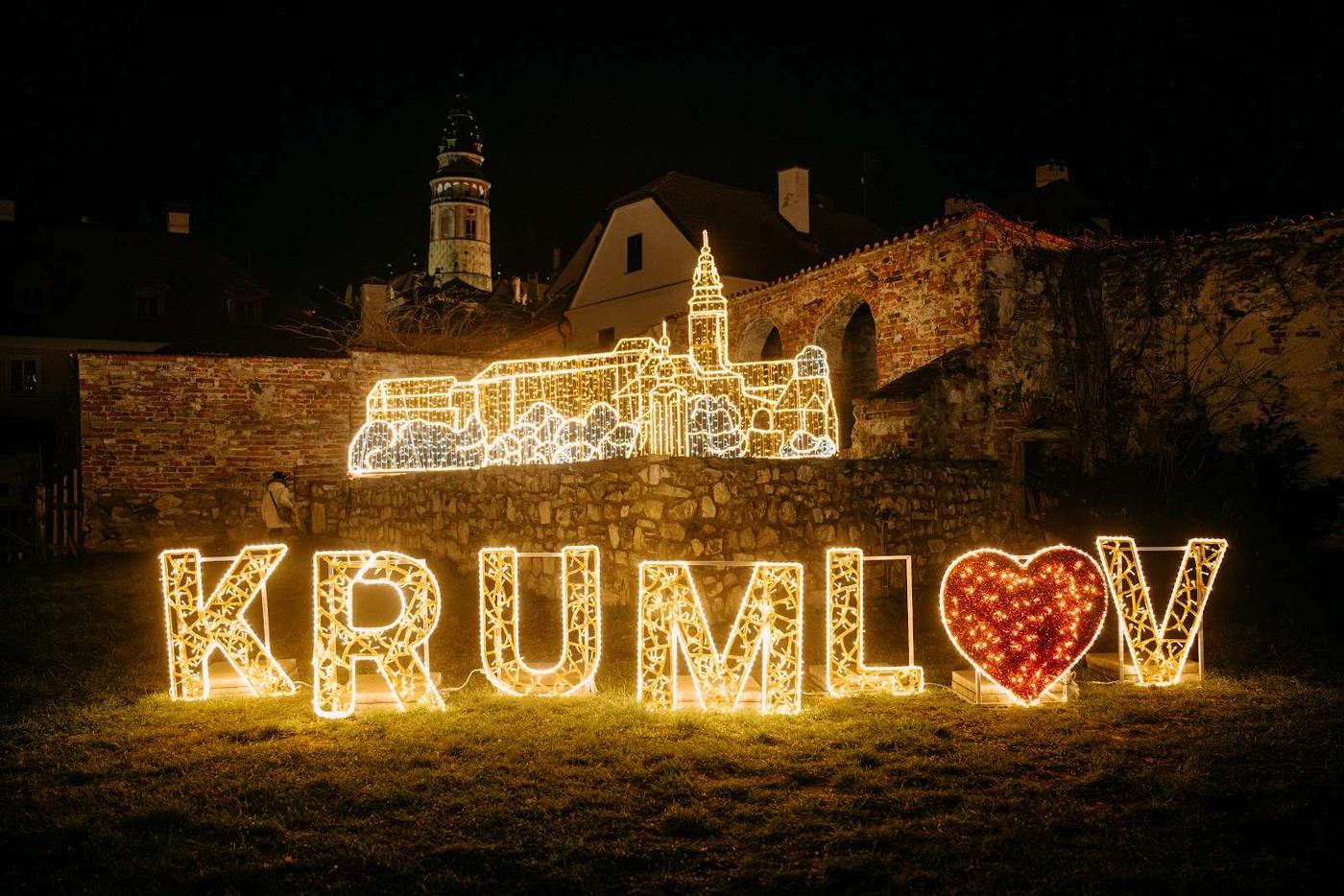 Světelný nápis KRUMLOV s červeným srdcem a silueta zámku před kamennou zdí v noci.